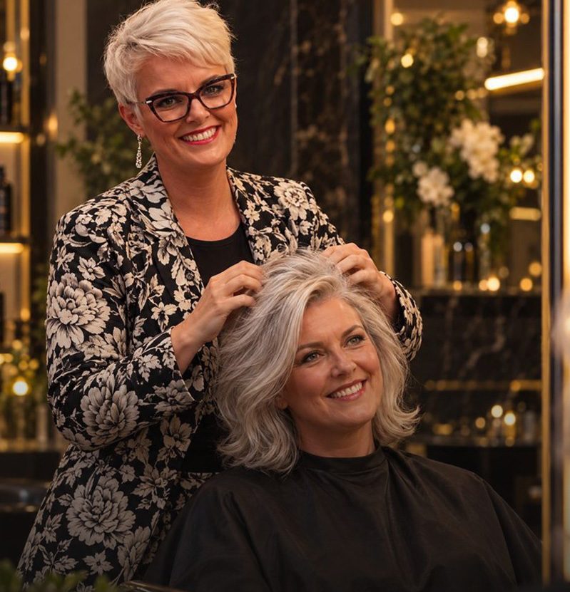 A stylist smiling in the mirror as her client’s silver hair is styled